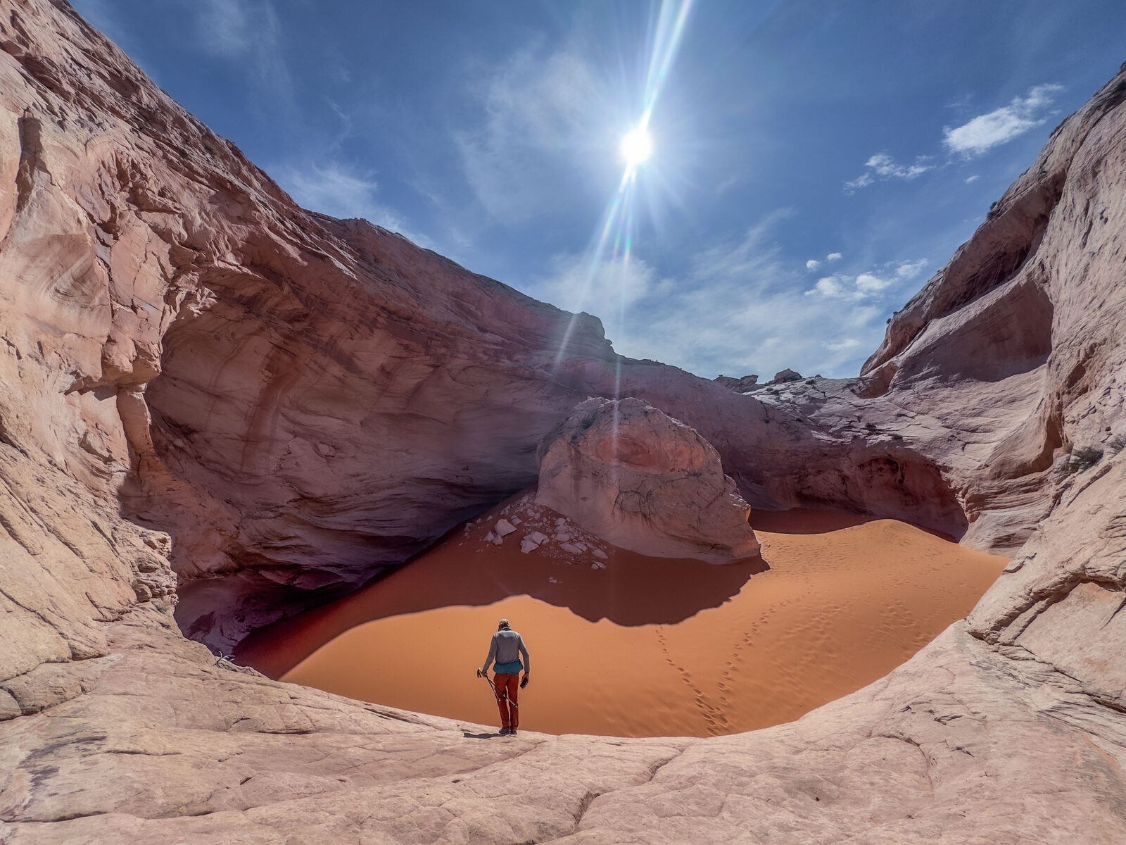 A lone hiker standing at the edge of the burnt-orange sand bowl, dwarfed by the slickrock walls and the sun overhead