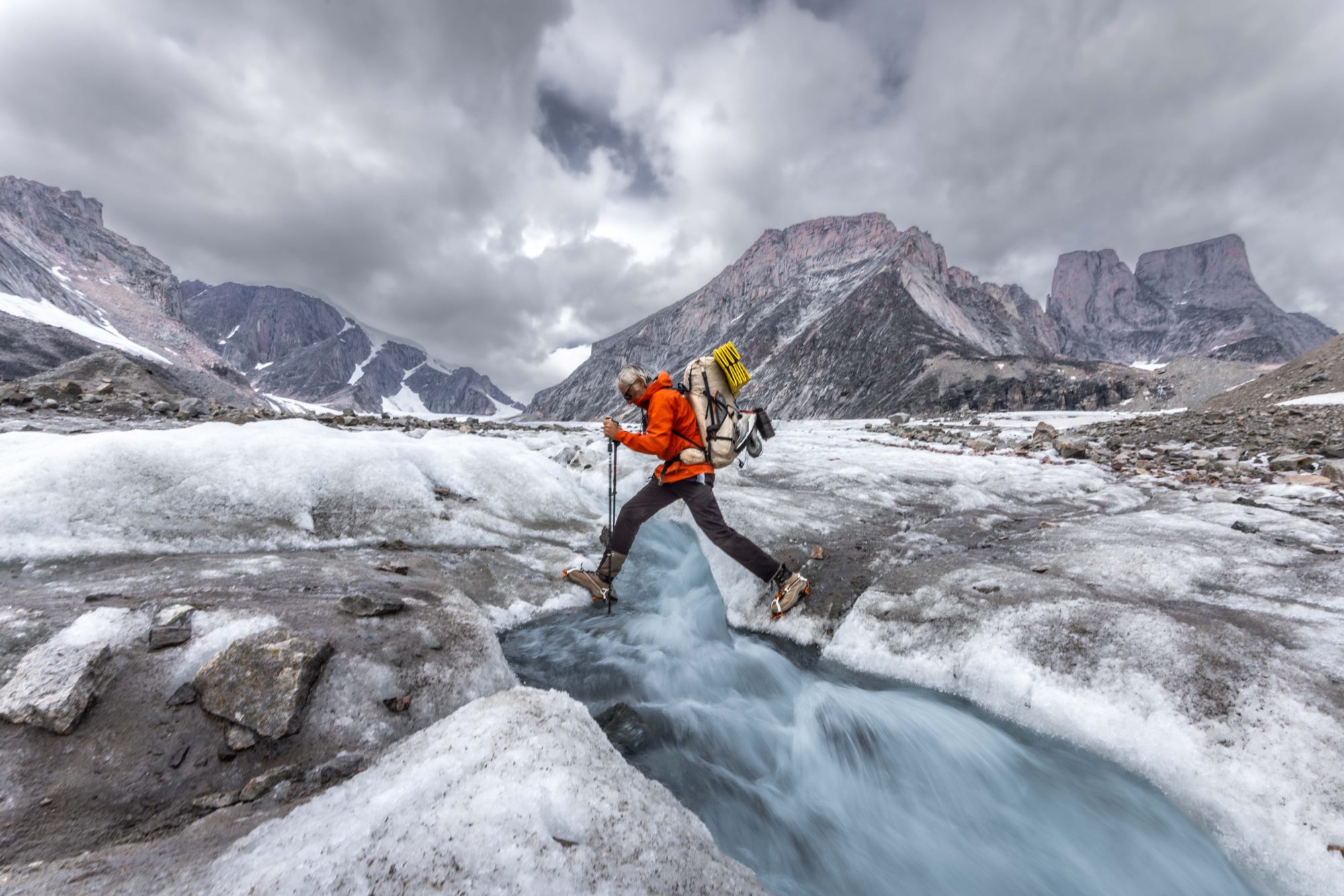 Paul Gagner leaping a glacial meltwater stream in Baffin Island with granite peaks and storm clouds behind him