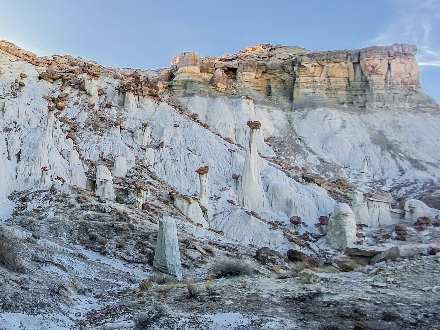 Wide view of the hoodoo field — a cliff wall above, white wash slope below, hoodoos stepping down through the middle